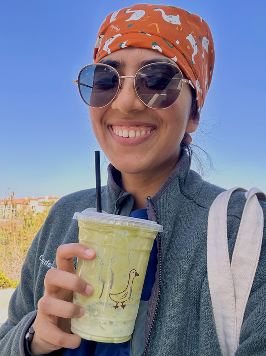 Person holding a drink with a straw, wearing sunglasses and a patterned scrub cap, against a clear blue sky.