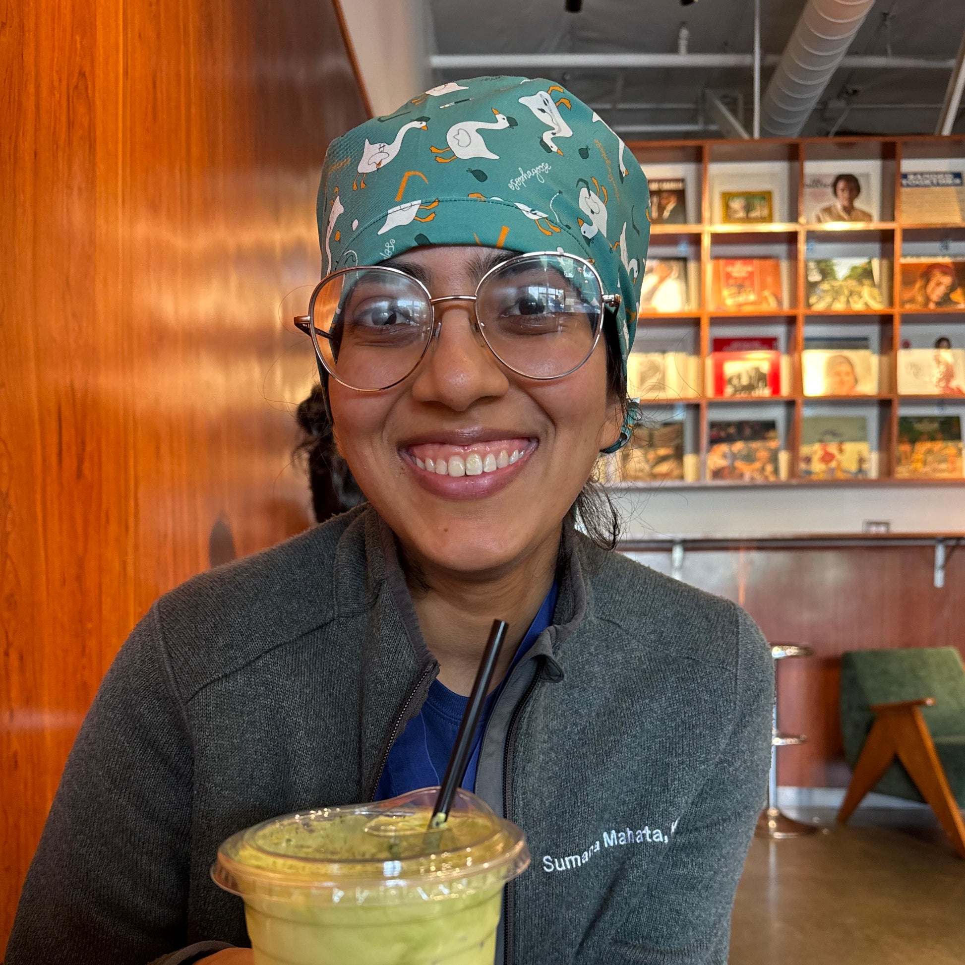 Person wearing a teal scrub cap with a goose pattern and holding a drink, sitting in a room with records in the background.