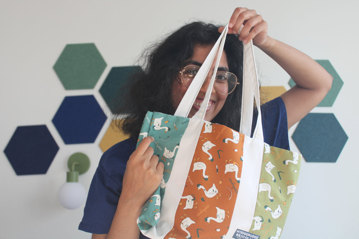 Hodgepodge holding up an esophagoose tote bag with three vertical panels featuring different goose prints in teal, orange, and green, against a white wall with hexagonal decals.