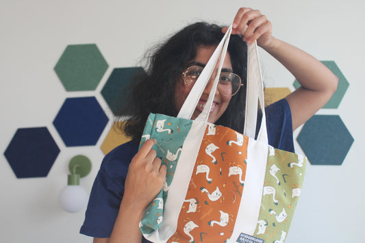 Hodgepodge holding up an esophagoose tote bag with three vertical panels featuring different goose prints in teal, orange, and green, against a white wall with hexagonal decals.