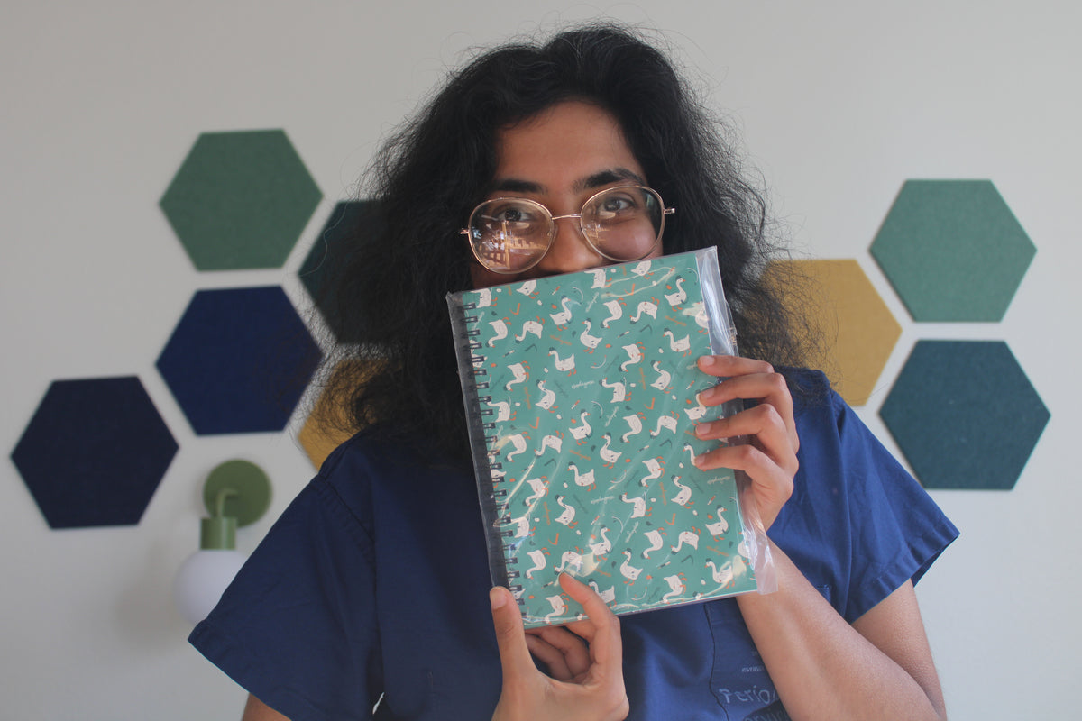 Hodgepodge holds an esophagoose in teal notebook in front of their face, wearing blue scrubs against a wall with green and yellow hexagonal panels.