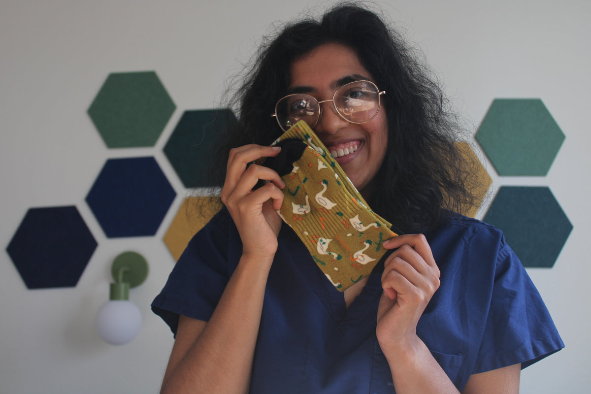 Hodgepodge smiles while holding an esophagoose in olive socks in front of her face. She is wearing blue scrubs, standing in front of a white wall decorated with green and navy hexagonal wall decals.