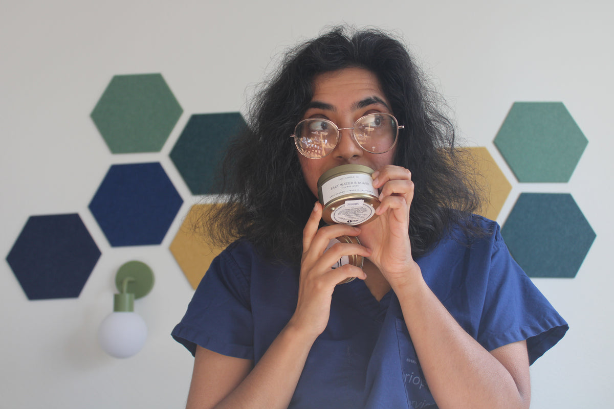 Hodgepodge holds the 1502 Candle to her nose, wearing blue scrubs in front of a wall with green and yellow hexagonal panels.