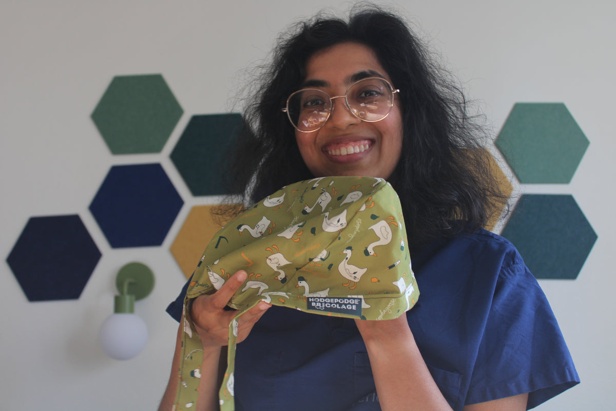 A smiling Hodgepodge with glasses and dark curly hair holds up an esophagoose scrub cap. She is wearing blue medical scrubs, standing in front of a white wall decorated with green and blue hexagonal wall decals.