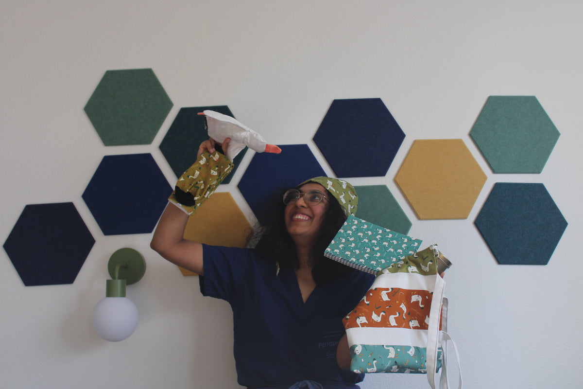 Hodgepodge in navy scrubs and the esophagoose in olive scrub cap holds the stuffed animal goose above her head while sitting on her couch trying to hold the esophagoose socks, notebook, tote bag, and candle. She is in a room with hexagonal wall panels in navy, green, and mustard yellow, with a green wall lamp on the left.