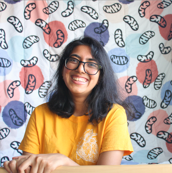 Sumana sitting in front of a mitochondria backdrop with a shirt that has an anatomic heart on it