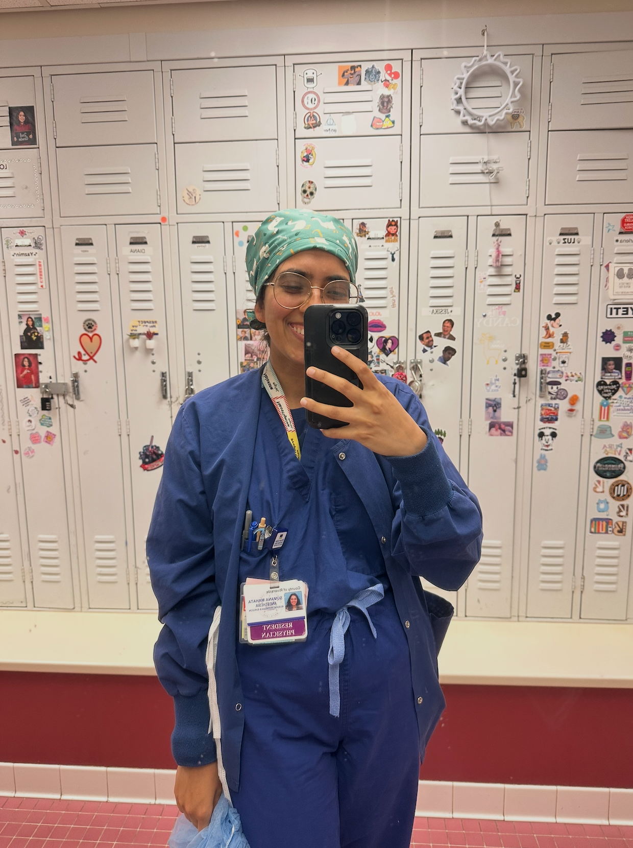 Person in blue scrubs taking a mirror selfie in front of lockers wearing the esophagoose scrub cap in teal.