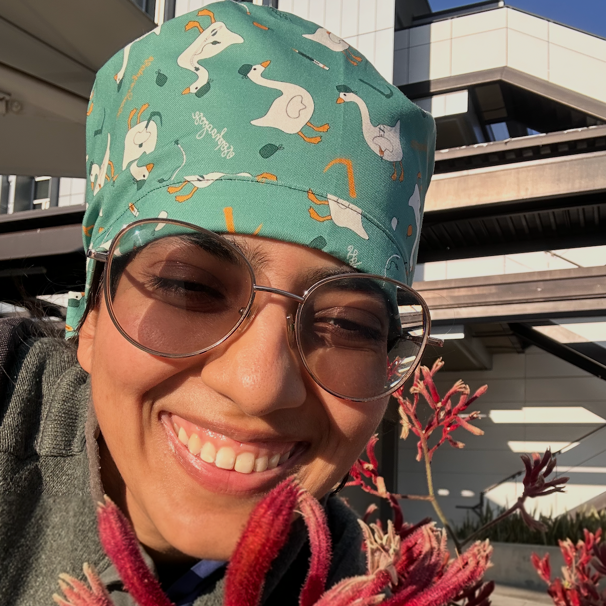 Person wearing a patterned scrub cap and glasses, standing outdoors with buildings and plants in the background.