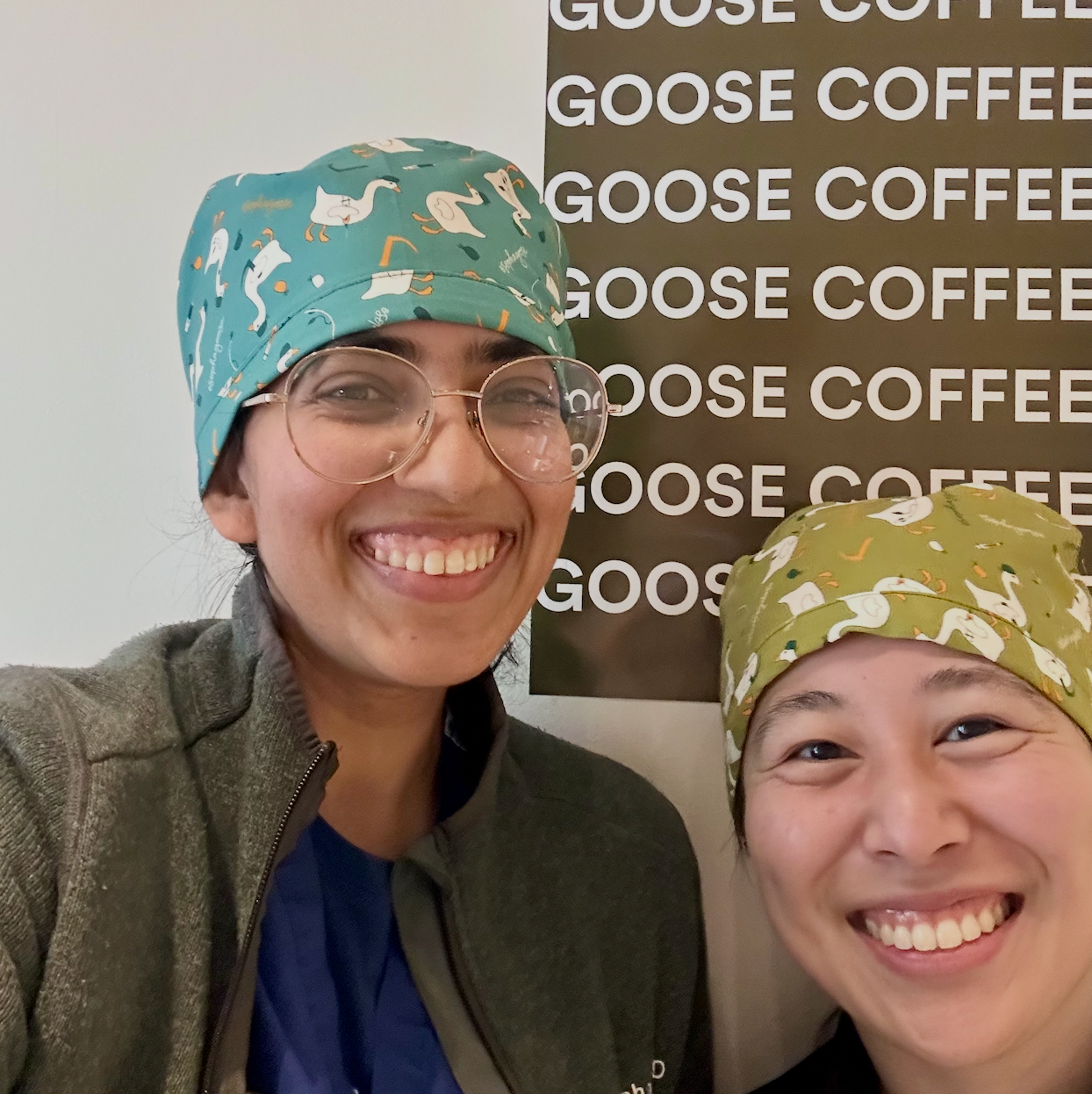 Two people wearing patterned scrub caps smiling in front of a 'Goose Coffee' sign.